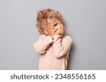 Adorable baby portrait. Shy baby girl with blonde wavy hair wearing rose shirt isolated over gray background hiding her face with arms looking through fingers to camera