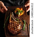 Small photo of Hands holding cooked Tomahawk (long bone ribeye) steak on a serving board. Low key image, vertical orientation