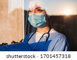 Young sad female caucasian UK US GP EMS doctor carer looking through ICU window,fear uncertainty in eyes,wearing face mask gazing at sun,hope faith in overcoming Coronavirus COVID-19 pandemic crisis 