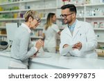Young male pharmacist giving prescription medications to senior female customer in a pharmacy with female pharmacist in the background