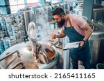 Man worker wearing an apron in a brewery using brewing equipment at factory. Control of plant, collecting data. Inspecting industrial equipment and making notes. Beer brewing process.