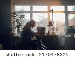 Small photo of Silhouette of a beautiful middle aged tailor woman sewing on a sewing machine while sitting at her moody working place at home. Fashion atelier, tailoring, handmade clothes concept. Slow Fashion.