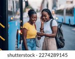 Two African american female friends waiting for a bus while at a bus stop and using a smartphone together. Riding, sightseeing, traveling to work, city tour, togetherness. Copy space.