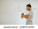 Small photo of Side view of a plasterer standing in a room in renovation process and using tools for plasterwork and skim coating walls. A worker is renovating house and doing manual building works. Copy space.