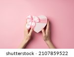 First person top view photo of valentine's day decorations young woman's hands holding heart shaped giftbox with pink ribbon bow on isolated pastel pink background