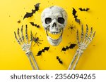 Embrace the eerie vibe of Halloween night's spookiness. Above view shot of a human skull and hands with bats and confetti on yellow isolated background, offering space for text or advertising content