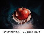 Small photo of Woman as witch offers red apple - symbol of toxic proposal, lure. Fairytale, white snow, wizard concept. Halloween celebration, cosplay. Smoke, mist background.