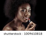 african american woman applying lipstick and looking at camera isolated on black