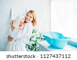 happy mother in bathrobe carrying adorable child covered in towel near plastic baby bathtub