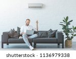 man turning on air conditioner with remote control while using laptop on sofa