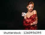 attractive girl in red shiny dress holding joker and queen of hearts cards isolated on black, looking at camera
