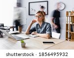 blonde businesswoman in eyeglasses sitting at workplace with gadgets in open space office, motion blur