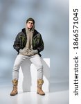 Small photo of full length of young trendy man in hat and anorak sitting with hands in pockets on white cube on grey