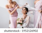 bridesmaids holding veil over pleased bride with wedding bouquet in bedroom