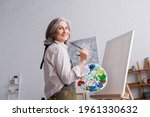 cheerful middle aged woman holding paintbrush and palette with colorful paints near blank canvas