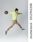 african american guy in hoodie, shorts and sneakers jumping with basketball on grey background