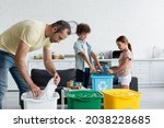 Man sorting garbage near blurred kids and trash cans with recycle sign at home