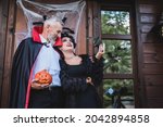 happy man in vampire costume holding carved pumpkin and embracing wife on cottage porch with halloween decoration