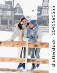 Small photo of full length of happy young woman in ear muffs looking at cheerful man in winter hat leaning on wooden border on ice rink
