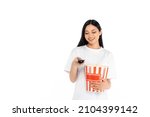 happy asian woman clicking tv channels while holding large popcorn bucket isolated on white
