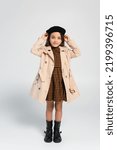 Small photo of full length of cheerful girl in stylish trench coat adjusting beret while standing on grey