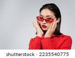 portrait of young asian woman adjusting red and stylish sunglasses while looking at camera isolated on grey
