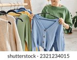 cropped view of tattooed woman with happy smile holding hanger with blue cardigan near rack with casual clothes at home, sustainable fashion and mindful consumerism concept