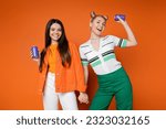 Cheerful blonde and brunette teenage girlfriends in casual outfits holding drink in tin cans and holding hands while standing on orange background, fashionable girls with sense of style