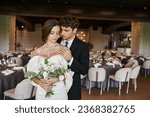 elegant groom with closed eyes embracing charming bride with wedding bouquet in modern event hall