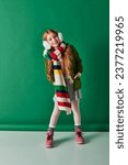 Small photo of full length, joyful girl in ear muffs, striped scarf and winter outfit on turquoise backdrop