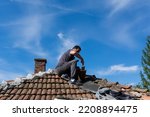 The man sitting on the roof of an old house and repairing tile with cement in the village on a sunny day. Blue sky in the background