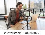 Cheerful man host with stubble laughing and gesticulating while streaming video podcast in broadcasting studio, using microphone, smartphone and laptop. Famous vlogger shooting video for his channel