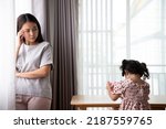 Small photo of Unhappy Asian mother touching forehead, feeling tired of bad daughter's behavior at home. Offended little child girl sitting on different side on table. stubborn and mischievous behavior