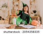 Small photo of cute little girl in a green Halloween costume of a witch or fairy prepares a pumpkin pie, barm brack in a festive decorated kitchen. space for text. High quality photo