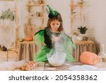 Small photo of cute little girl in a green Halloween costume of a witch or fairy prepares a pumpkin pie, barm brack in a festive decorated kitchen. space for text. High quality photo