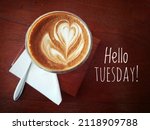 Small photo of Tuesday coffee with text greeting on brown wooden table - Hello Tuesday. With cup of coffee latte and spoon. Happy Tuesday.