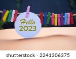 Hello 2023. New Year 2023 concept with golden text on white circle paper and colorful wooden clips on rope on dark and light orange background. Fresh hello 2023. Happy New Year.