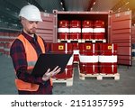 Logistician in warehouse of oil products. Barrels of fuel next to man. Male logistician standing working on laptop. Sea container is filled with barrels of oil. Preparation of oil for export.
