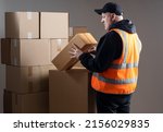 Small photo of Postman looks at box. Postman in orange vest on gray. Postal service worker with boxes. Mature postman back to camera. Career in post office concept. Man in black baseball cap in warehouse