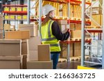 Small photo of Bonded warehouse. Woman customs officer holding boxes. Girl in yellow vest with her back to camera. Boxes and tiered racks. Customs warehouse with cardboard boxes. Career customs worker.