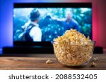 A glass bowl of popcorn and remote control in the background the TV works. Evening cozy watching a movie or TV series at home.