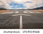 White arrow on a small asphalt air plane runway. Cloudy sky with small blue opening in the middle. Sun rays and flare. Small airport on the right. Aviation industry theme. Selective focus