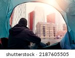 View from inside of a homeless tent. Man in dark dirty cloths sitting by the entrance looking at rich high value district houses. Living during financial crisis concept. Dreaming on better future.