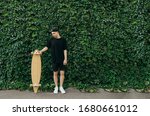 Young man in dark clothing and a cap stands on the background of an ivy wall, holding a longboard in his hand. Guy with a longboard poses on a background of green wall with ivy.Background