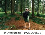 Young male hiker with a backpack stands in a mountain untouched forest and takes a photo on a smartphone camera, rear view. Guy on the hike uses a smartphone camera.