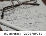 Small photo of Mathematical equations written on a piece of paper containing cosines, sines, square roots, fractions. Glasses and a pen are on the sheet. Desktop.