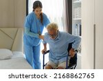Small photo of Asian smiling nurse helping senior man get out of bed nursing care support patient while getting out of bed and moving to wheelchair at home helping handicapped elderly stand up