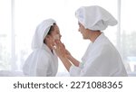 Small photo of Asian mother adding treatment cream on the cheek to little girl with spa dress and head covered with a white towel on bed together at home