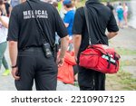 Medical first responders walk along a road wearing black uniforms, with medical first responder in grey letters across the back of the paramedic. The EMT is carrying a red first aid kit and radio.