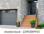 Small photo of A grey colored modern garage door, two large flower pots with mums and cascading flowers at the end of brown wooden stairs to a black panel door. The exterior wall of the house is grey colored brick.